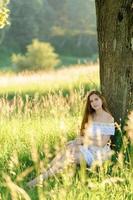 portret van een jong mooi meisje in een zomerjurk. zomer fotosessie in het park bij zonsondergang. een meisje zit onder een boom in de schaduw. foto