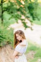 portret van een jong mooi meisje in een zomerjurk. zomer fotosessie in het park bij zonsondergang. een meisje zit onder een boom in de schaduw. foto