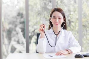 professionele aziatische vrouwelijke arts draagt een medische jas en stethoscoop in de kantoorruimte terwijl hij naar de camera in het ziekenhuis kijkt met het concept van de gezondheidscontrole. foto