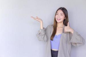 mooie jonge aziatische vrouw die een paars shirt draagt, handelt duim omhoog als een goed symbool en een andere hand toont iets op de achtergrond. foto