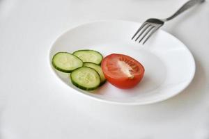 hele en gesneden komkommer en tomaat op een witte plaat en vork foto