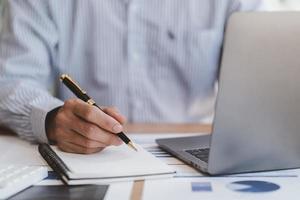 zakenmanhand maakt aantekeningen op papier met een zwarte pen en gebruikt een laptopcomputer op een wit bureau op kantoor. foto