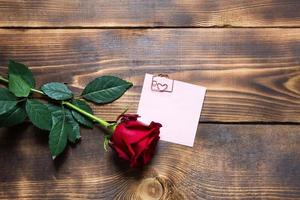 rode roos op houten achtergrond met copyspace en sticker met een paperclip en hart, een herinnering op een notitieblad. cadeau voor vrouw op vakantie, een uitnodiging voor een date, valentijnsdag, een teken van liefde foto
