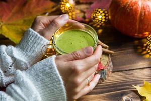 groene japanse matcha-thee met schuim in transparante beker op houten tafel in het herfststilleven. vrouwenhand met lange witte truimouwen wordt verwarmd door een drankje. focus op beker. warme sfeer en comfort foto