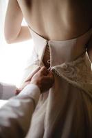 prachtige, blonde bruid in een witte luxe jurk die zich klaarmaakt voor de bruiloft. ochtend voorbereidingen. een vrouw trekt een jurk aan. de bruidegom helpt de bruid. foto