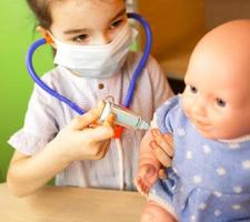 een meisje met een pop speelt dokter, maakt een injectiespuit in de hand. vaccinatie, vaccinatiekalender, vaccin, beroepspel. injectie van een verpleegster foto