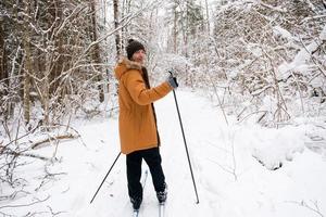 skiër in hoed met pompon met skistokken in zijn handen met zijn rug tegen de achtergrond van een besneeuwd bos. langlaufen in het winterbos, buitensporten, gezonde levensstijl. foto