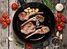 geroosterde lamsribben met rozemarijn, tomaten en knoflook op pan op donkere oude rustieke houten achtergrond, bovenaanzicht foto