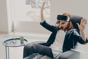 jonge opgewonden roodharige vrouw die een vr-bril of -bril test terwijl ze thuis op een fauteuil zit foto