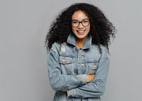 lachende gekrulde vrouw met zachte glimlach, in een goed humeur, draagt een bril, houdt de armen over elkaar, draagt denim kleding, kijkt recht naar de camera, geniet van een leuk gesprek, geïsoleerd op een grijze achtergrond foto