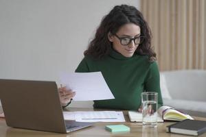 jonge, aangename Italiaanse vrouw met een bril die een rapport voorbereidt terwijl ze op een laptop in een modern kantoor werkt foto