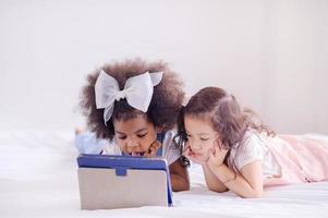een half-Afrikaans meisje en haar Aziatische vriendin liggen in bed en kijken naar online leerprogramma's voor kinderen foto