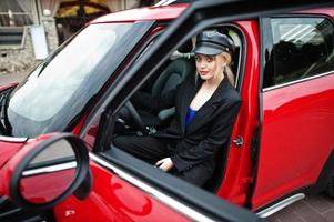 portret van mooie blonde sexy mode vrouw model in pet en in alle zwart met lichte make-up zitten en rijden rode stadsauto. foto