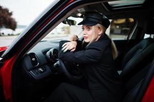 portret van mooie blonde sexy mode vrouw model in pet en in alle zwart met lichte make-up zitten en rijden rode stadsauto. foto