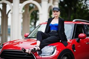 portret van mooie blonde sexy mode vrouw model in pet en in alle zwart met lichte make-up zitten op de motorkap van de rode stadsauto. foto
