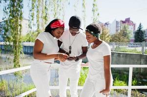 drie stijlvolle en trendy Afro-Amerikaanse vrienden, draag witte kleding. straatmode van jonge zwarte mensen. zwarte man met twee Afrikaanse meisjes die op de telefoon kijken. foto