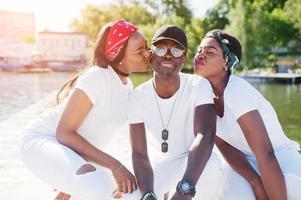 drie stijlvolle Afro-Amerikaanse vrienden, draag witte kleren op de pier op het strand. straatmode van jonge zwarte mensen. zwarte man met twee Afrikaanse meisjes kuste hem en liet lippen zien. foto