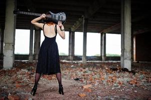 roodharige punkmeisjeslijtage op zwart met basgitaar op verlaten plaats. portret van gotische vrouw muzikant. foto