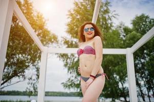 portret van een aantrekkelijk meisje poseren in bikini met zonnebril op terwijl je op een wit houten terras staat. foto