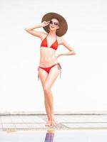 sexy meisje gelukkig meisje in rode bikini dragen hoed en zonnebril in de buurt van zwembad met witte muur. foto