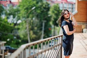 brunette meisje op zwarte jurk, zonnebril poseren op straat van de stad. foto