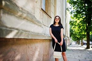 brunette meisje op zwarte jurk op zonnebril met handtas bij de hand poseren op straat van de stad. foto