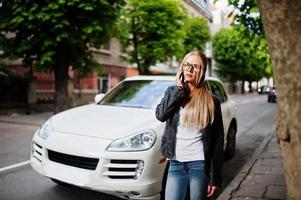 stijlvolle blonde vrouw draagt jeans, bril en leren jas met mobiele telefoon, tegen luxe auto. mode stedelijk model portret. foto