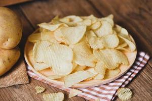 chips snack op wit bord, krokante chips op de keukentafel en verse rauwe aardappelen foto