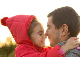klein meisje in een rood jasje met een capuchon knuffelt en kust haar vader, glimlacht, raakt haar neus aan. gelukkige familie, emoties van kinderen, vaderdag, felle zonnestralen, kaukasische uitstraling. ruimte voor tekst. foto