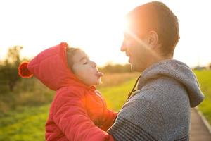 klein meisje in een rood jasje met een capuchon knuffelt en steekt tong uit haar vader, glimlacht. gelukkige familie, emoties van kinderen, vaderdag, felle zonnestralen, kaukasische uitstraling. ruimte voor tekst. foto