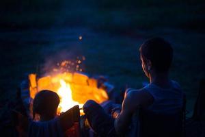 vader en dochter zitten 's nachts bij het vuur in de open lucht in de zomer in de natuur. familie camping trip, bijeenkomsten rond het kampvuur. vaderdag, barbecue. camping lantaarn en tent foto