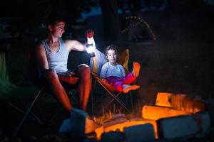 vader en dochter zitten 's nachts bij het vuur in de open lucht in de zomer in de natuur. familie camping trip, bijeenkomsten rond het kampvuur. vaderdag, barbecue. camping lantaarn en tent foto