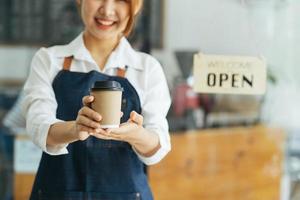 portret van een gelukkige vrouw die bij de deuropening van haar winkel staat. vrolijke volwassen serveerster wachten op klanten bij coffeeshop. succesvolle eigenaar van een klein bedrijf in casual gekleed in een blauwe schort die bij de ingang staat foto