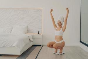 portret van opgewonden jonge, mooie blanke vrouw hurkt op een weegschaal in de slaapkamer foto