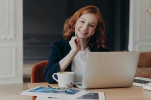 freelance zakenvrouw met rood haar zit voor een laptopcomputer, communiceert met collega's via videoconferentie, zit op het bureaublad, drinkt koffie, heeft een vrolijke uitdrukking. afstand baan foto