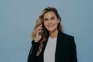 zelfverzekerde positieve glimlachende jonge vrouwelijke kantoormedewerker die op smartphone praat foto