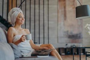 foto van ontspannen lachende vrouw drinkt graag thee, leest een tijdschrift en poseert op een comfortabele bank in de woonkamer, geniet van vrije tijd tijdens de vrije dag, draagt minimale make-up, heeft een goed verzorgde, gezonde huid