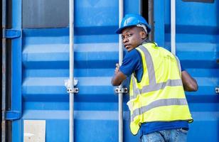 slimme afro-amerikaanse ingenieur man met industriële werkkleding overall en beschermende helm die voorraadcontrole doet en de deur van de zware container in de containerwerf op de magazijnachtergrond sluit foto