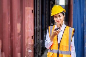 jonge zelfverzekerde vrouwelijke ingenieur glimlacht en gebruikt radiocommunicatie en draagt een gele veiligheidshelm en controleert op controle laadcontainers doos van vrachtvrachtschip voor import en export foto