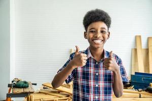 lachende Afro-Amerikaanse jongen timmerman permanent met duimen opgevend als teken van succes in een timmerwerkplaats. foto