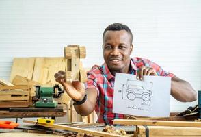 jonge afro-amerikaanse mannelijke timmerman zittend aan tafel met modelauto houten en tekenpapier via tablet terwijl video online chatten met klant of online lesgeven in de fabriek foto