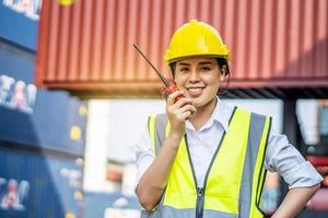 jonge zelfverzekerde vrouwelijke ingenieur glimlacht en gebruikt radiocommunicatie en draagt een gele veiligheidshelm en controleert op controle laadcontainers doos van vrachtvrachtschip voor import en export foto