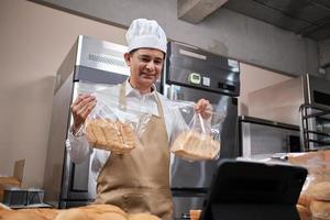 senior Aziatische mannelijke chef-kok in schort, blogger voor kleine bedrijven livestreaming via mobiele applicatietechnologie, online promotie van bakkerijverkoop, vers brood, banketbakkerswaren en commercieel koken in de keuken. foto