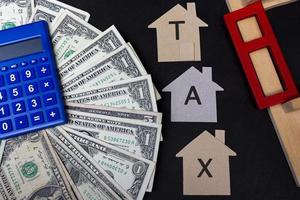 belastingtekst op bruin papieren huismodel met dollarbankbiljetten, blauwe rekenmachine en raamkozijn op donkere houten tafel. concept voor onroerendgoedbelasting. foto
