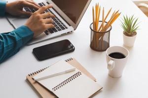 jonge man met laptopcomputer en mobiele telefoon bij het zoeken naar financiële informatie in het bedrijfsleven, werk aan de balie. schrijven met een pen, op afstand studeren vanuit huis en thuiswerken. foto