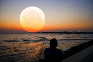 man in silhouet kijkend naar de afstand naar de kalme oranje volle maan overzee met een paars fluweel en kalme oranje lucht. foto