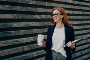 blije roodharige jonge vrouw drinkt afhaalkoffie houdt digitale tablet en notitieblok vast foto