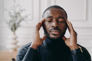 gestresste Afrikaanse thuiskantoormedewerker zit aan het bureau en raakt tempels aan terwijl de ogen gesloten zijn foto