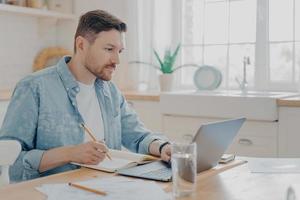 geconcentreerde jonge zakenman die notities schrijft over de agenda en op afstand werkt op laptop foto