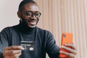 Glimlachende Afro-Amerikaanse zakenman die creditcard en smartphone gebruikt terwijl hij op de werkplek zit foto